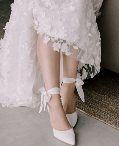 White pointed-toe heels made of leather, featuring delicate ankle ties. The shoes are worn by a person whose legs are visible, adorned with a floral-embroidered white dress. The image is taken from a low angle, showcasing the shoes against a neutral background with a textured rug.