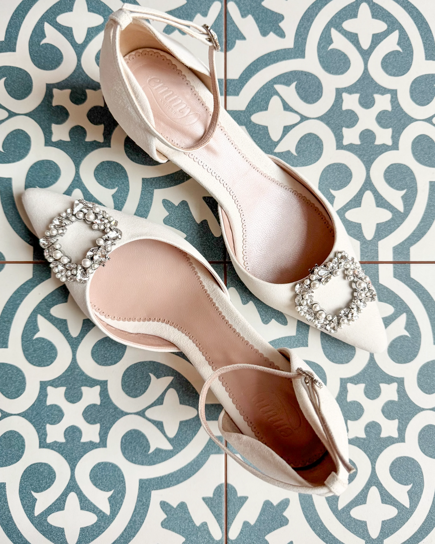 Elegant white shoes featuring a decorative crystal buckle on the toe. The shoes are positioned on a patterned tiled surface, showcasing their pointed toe design. The angle captures the shoes from above, highlighting the intricate details of the buckle and the smooth finish of the material.