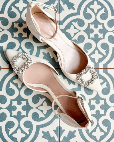 Elegant white shoes featuring a decorative crystal buckle on the toe. The shoes are positioned on a patterned tiled surface, showcasing their pointed toe design. The angle captures the shoes from above, highlighting the intricate details of the buckle and the smooth finish of the material.
