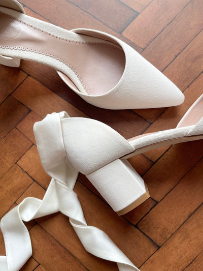 White suede heels with a square block heel, approximately 3 inches in height. The shoes are positioned on a wooden floor, showcasing their pointed toe design. A white satin ribbon is elegantly draped beside the heels, adding a touch of sophistication to the overall presentation.